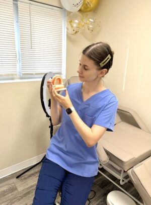 A woman in blue scrubs sits in a medical office, holding a dental model of a mouth. She looks at the model attentively, with balloons and a ring light in the background.