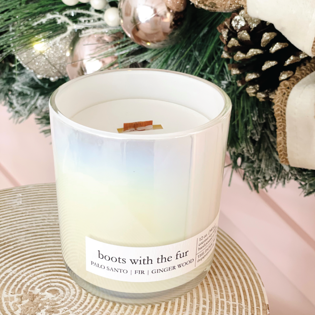 A white candle labeled "boots with the fur" sits on a textured surface in front of a festive holiday wreath with pinecones, ornaments, and ribbon.