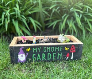A wooden planter box labeled "My Gnome Garden" with painted images of a gnome and mushrooms, containing small gnome and frog figurines, sits on grass with green plants in the background.
