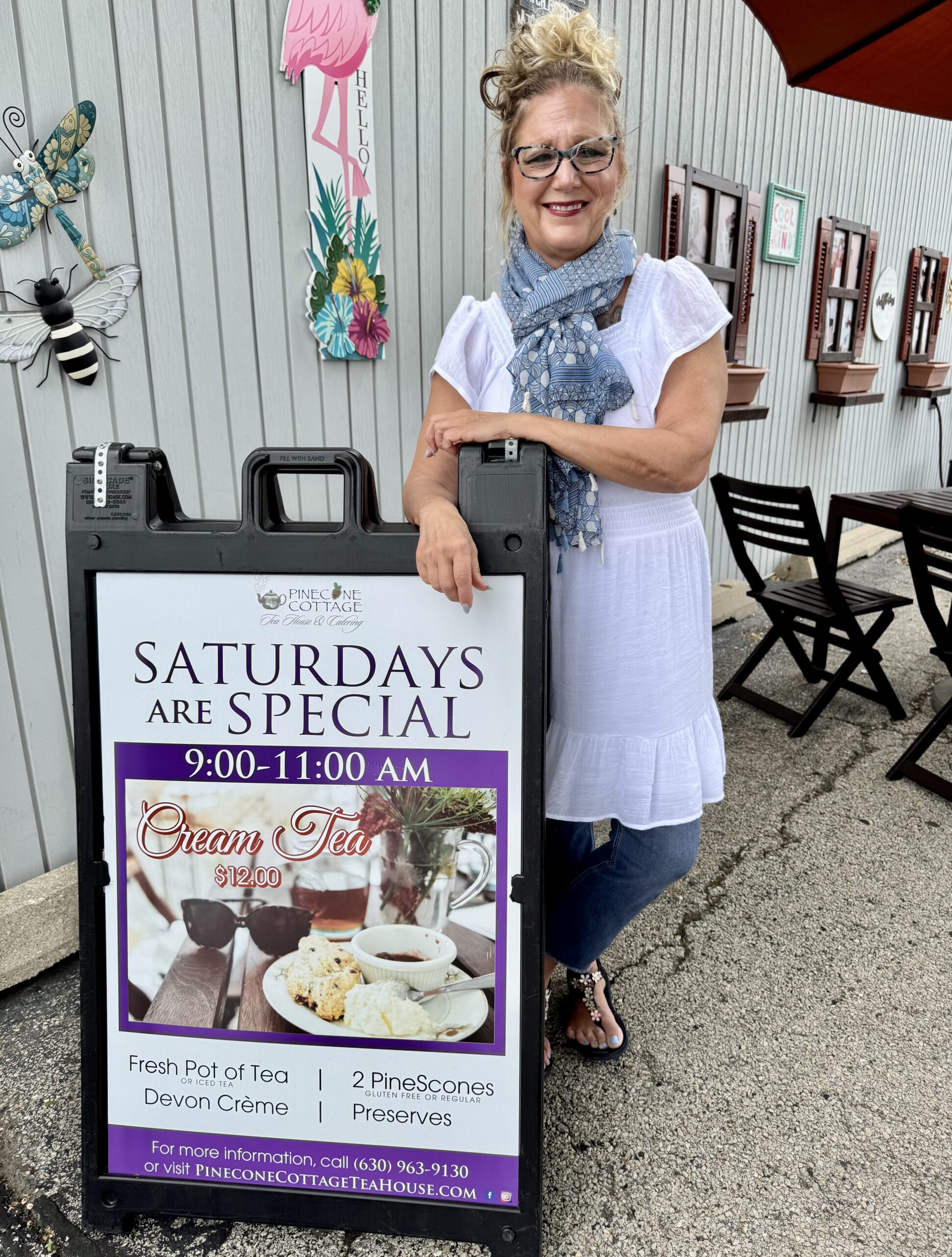 A smiling woman in a white dress and blue scarf stands next to a sign advertising "Saturdays are Special: Cream Tea, 9–11 AM" with tea, scones, and preserves at Pinecone Cottage Tea House.