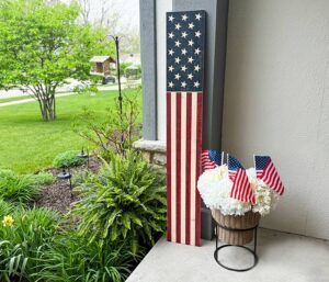 A tall wooden decoration painted like the American flag leans against a porch wall next to a basket of white flowers with small American flags, surrounded by green plants and a well-kept lawn.
